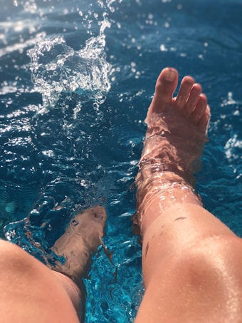 A pair of legs are submerged in a pool, creating splashes and ripples in the clear blue water. Sunlight glistens on the skin and water, adding a sense of warmth and relaxation.