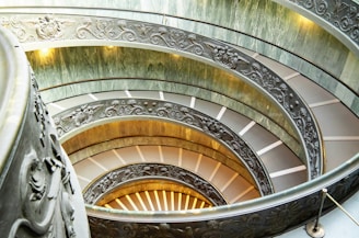 An intricate spiral staircase with ornate metal railings featuring decorative patterns. The staircase curves upward, creating a dynamic and elegant visual effect. Soft lighting enhances the ambiance, highlighting the detailed craftsmanship of the designs.
