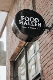 A circular black and white sign with the words 'Food Hallen Rotterdam' is mounted on the brick wall of a building. The wall features large windows that reflect a nearby structure.