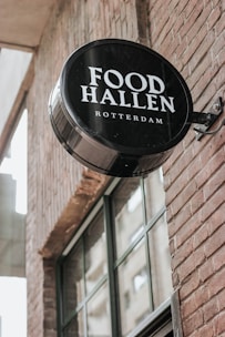 A circular black and white sign with the words 'Food Hallen Rotterdam' is mounted on the brick wall of a building. The wall features large windows that reflect a nearby structure.