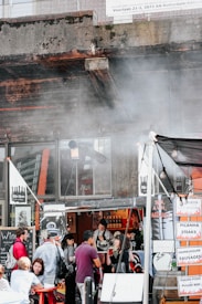 A small, lively street food stall under a gritty urban overpass with people gathered around. Signs advertising food items like 'Picanha Steaks' and 'Pulled Pork' are visible. The environment is bustling, with patrons chatting and enjoying beverages. Smoke emanates from the stall, adding an authentic street food ambiance.