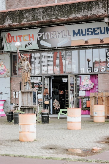 Front view of a building with a sign indicating 'Dutch Pinball Museum.' The entrance is open, revealing some pinball machines inside. Two people are seen entering or exiting the museum. Various elements including a statue holding a light, colorful drums, and outdoor furniture such as a bench and table are visible outside.