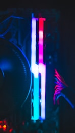 A close-up view of computer components inside a case with vibrant RGB lighting. Two RAM sticks are illuminated with a gradient of blue, pink, and red colors. Cables with a red accent are visible to the right, contributing to the colorful and high-tech appearance.