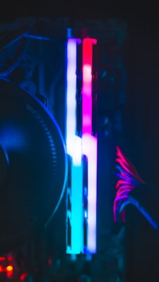 A close-up view of computer components inside a case with vibrant RGB lighting. Two RAM sticks are illuminated with a gradient of blue, pink, and red colors. Cables with a red accent are visible to the right, contributing to the colorful and high-tech appearance.