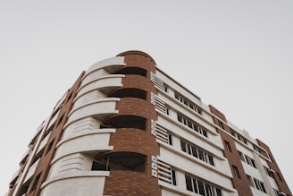 A modern multi-story building with a combination of red brick and white concrete. The structure features alternating curved balconies and rectangular windows, creating an architectural pattern. The sky is overcast and provides a neutral backdrop.