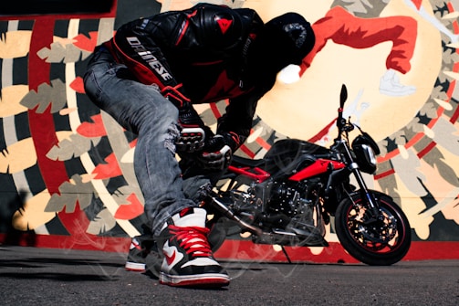 A person wearing a black and red motorcycle jacket and ripped jeans is kneeling in front of a red and black motorcycle. The background features a vibrant mural with abstract patterns and a large depiction of a person in red. The person is wearing red and white sneakers and appears to be engaging with the motorcycle.