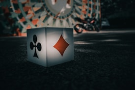 A large cube with black club and red diamond symbols on its sides is placed on the ground. In the blurred background, there is a colorful, circular pattern on a wall and a motorcycle parked next to it. The scene is bathed in soft, shadow-dappled light, creating a mysterious ambiance.
