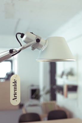 A modern desk lamp with a minimalist design is prominently displayed in an office space. The lamp's arm and head are silver, and a tag with the brand name 'Artemide' is attached to it. The background features blurred office furniture and greenery, creating a soft-focus backdrop.