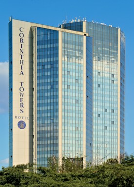 A tall modern building with a glass facade reflecting the sky, featuring the name 'Corinthia Towers Hotel' prominently displayed. The structure is set against a clear blue sky and surrounded by some greenery at the base.
