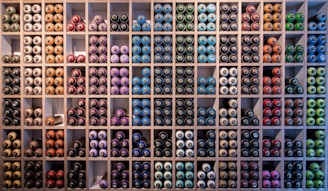 A neatly organized wall filled with rows of spray paint cans, arranged by colors such as blue, green, red, purple, and orange. The cans are stored in individual square compartments, creating a vibrant and orderly display.