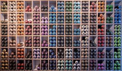 A neatly organized wall filled with rows of spray paint cans, arranged by colors such as blue, green, red, purple, and orange. The cans are stored in individual square compartments, creating a vibrant and orderly display.