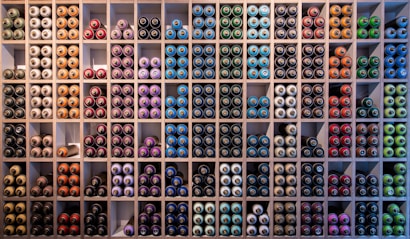 A neatly organized wall filled with rows of spray paint cans, arranged by colors such as blue, green, red, purple, and orange. The cans are stored in individual square compartments, creating a vibrant and orderly display.