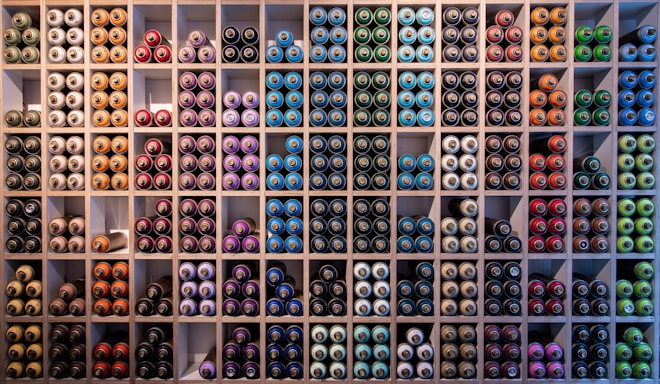 A neatly organized wall filled with rows of spray paint cans, arranged by colors such as blue, green, red, purple, and orange. The cans are stored in individual square compartments, creating a vibrant and orderly display.