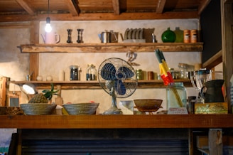 A rustic kitchen setting features wooden shelves adorned with various jars, bottles, and pottery. A vintage blue fan, a pineapple, woven baskets, and a jar with colorful utensils are placed on the wooden counter. A pendant light bulb hangs from the ceiling, casting warm light throughout the scene.