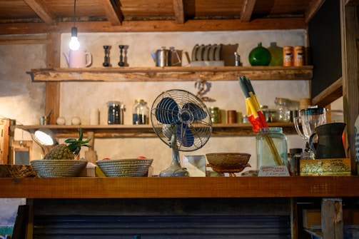 A rustic kitchen setting features wooden shelves adorned with various jars, bottles, and pottery. A vintage blue fan, a pineapple, woven baskets, and a jar with colorful utensils are placed on the wooden counter. A pendant light bulb hangs from the ceiling, casting warm light throughout the scene.
