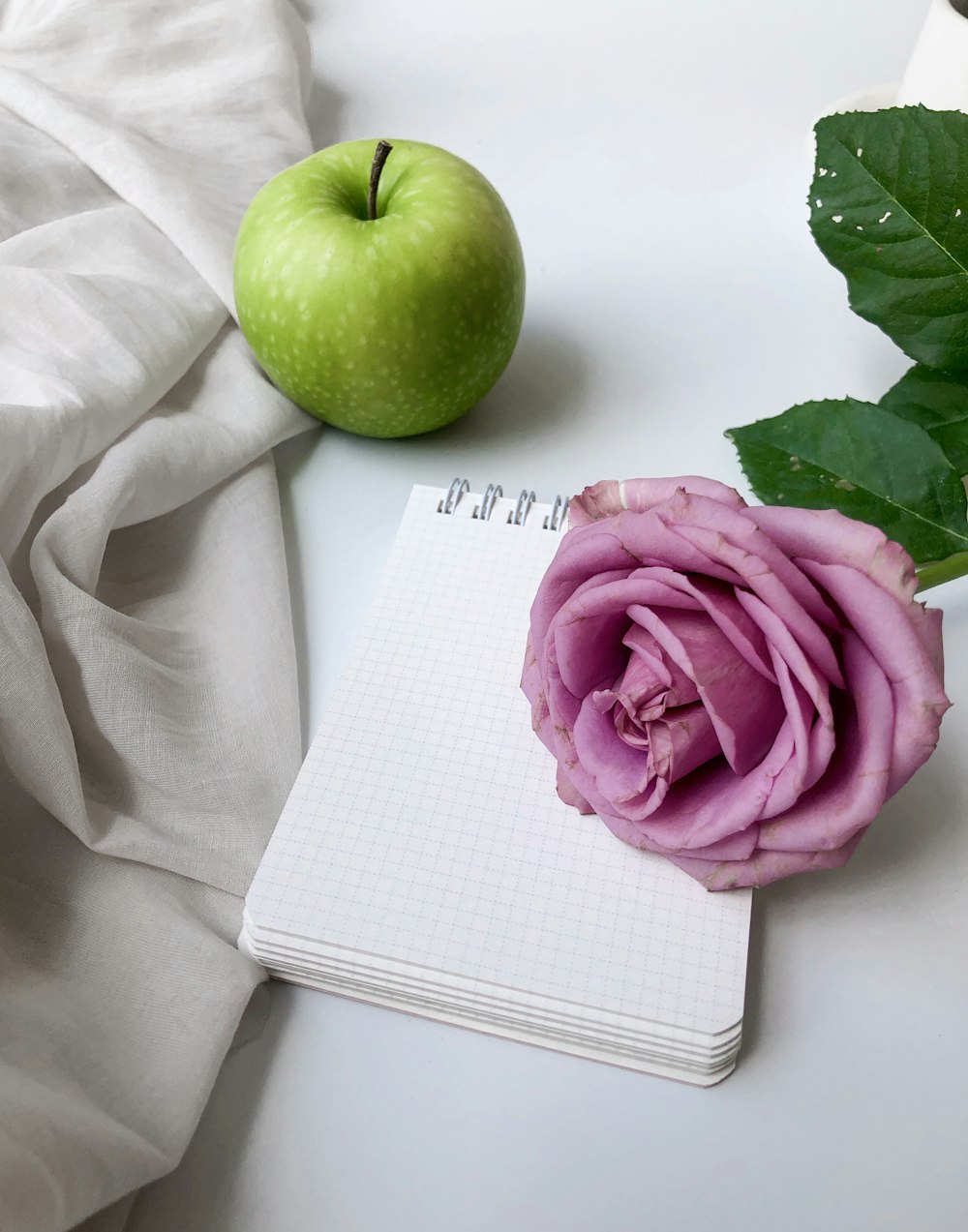 green apple near a pink petaled rose flower and white mini-notebook