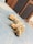 A piece of turmeric root lies on a dusty blue surface. The root appears fresh, with visible orange flesh and dry, dusty brown skin. Thin, dry root strands extend from the main body, and the background features a wooden texture partially visible.