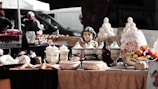 A rustic market stall displays a collection of decorative items including ceramic pots, vases, and jars with ornate designs. At the center, a cherubic statue with wings is prominently featured. The stall also includes a variety of dried goods and fruits arranged artfully. In the background, people are casually walking past other stalls, and several vehicles are parked.