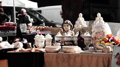 A rustic market stall displays a collection of decorative items including ceramic pots, vases, and jars with ornate designs. At the center, a cherubic statue with wings is prominently featured. The stall also includes a variety of dried goods and fruits arranged artfully. In the background, people are casually walking past other stalls, and several vehicles are parked.