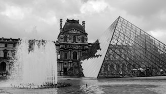 A black and white photograph featuring a large glass pyramid structure next to an ornate historical building, with an active water fountain in the foreground. The sky is overcast with thick clouds.