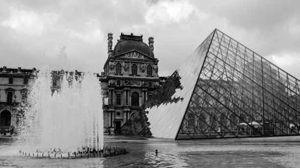 A black and white photograph featuring a large glass pyramid structure next to an ornate historical building, with an active water fountain in the foreground. The sky is overcast with thick clouds.