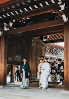 A group of people dressed in traditional Japanese attire are walking under a wooden gate with intricate designs. The primary focus is on a person in white formal clothing, possibly a wedding attire, accompanied by others in kimonos. The background includes elements of traditional Japanese architecture, with another group of people visible through the gate.