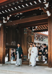 A group of people dressed in traditional Japanese attire are walking under a wooden gate with intricate designs. The primary focus is on a person in white formal clothing, possibly a wedding attire, accompanied by others in kimonos. The background includes elements of traditional Japanese architecture, with another group of people visible through the gate.