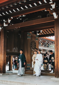 A group of people dressed in traditional Japanese attire are walking under a wooden gate with intricate designs. The primary focus is on a person in white formal clothing, possibly a wedding attire, accompanied by others in kimonos. The background includes elements of traditional Japanese architecture, with another group of people visible through the gate.