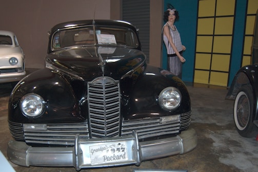 A vintage black car, prominently displayed with a license plate reading 'Grandpa's 1947 Packard', is flanked by another classic white car on the left. A mannequin in period clothing stands in the background near a yellow and blue wall.