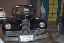 A vintage black car, prominently displayed with a license plate reading 'Grandpa's 1947 Packard', is flanked by another classic white car on the left. A mannequin in period clothing stands in the background near a yellow and blue wall.