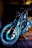 A motorcycle with a metallic finish is parked indoors, showcasing intricate details of its wheel and engine components. The lighting emphasizes the chrome and mechanical features, casting a cool, dramatic illumination on the bike and floor.
