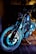 A motorcycle with a metallic finish is parked indoors, showcasing intricate details of its wheel and engine components. The lighting emphasizes the chrome and mechanical features, casting a cool, dramatic illumination on the bike and floor.