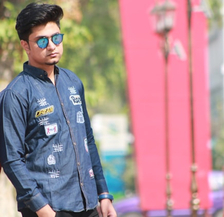 A person wearing a denim shirt adorned with patches and embroidery stands outdoors. They are sporting reflective sunglasses and have their hands in their pockets. In the background, there is a blurred view of trees and a large red banner or structure, contributing to a casual and relaxed atmosphere.