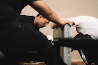 Two individuals are leaning against a concrete bench in an indoor setting, appearing focused and engaged. One person is wearing glasses and has short hair.