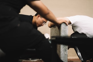 Two individuals are leaning against a concrete bench in an indoor setting, appearing focused and engaged. One person is wearing glasses and has short hair.