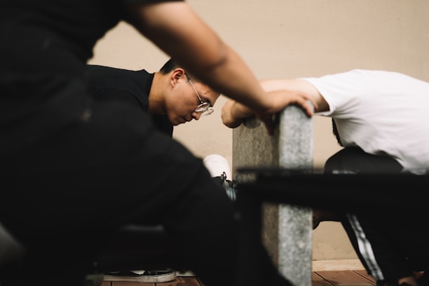 Two individuals are leaning against a concrete bench in an indoor setting, appearing focused and engaged. One person is wearing glasses and has short hair.