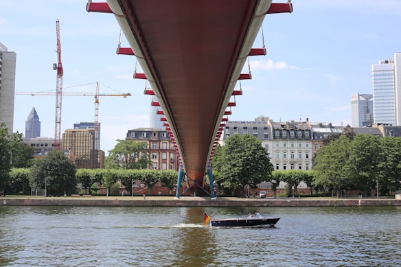 A sleek boat with a German flag moves across a river under a large, modern bridge. On the opposite riverbank, there are several tall buildings and construction cranes towering over older, more traditional European-style buildings and greenery.