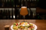 A tall glass of beer on a wooden table, accompanied by a salad on a white plate and a small shot glass with brown liquid. The background is dimly lit, with blurred reflections and shadows, suggesting a cozy and relaxed atmosphere.