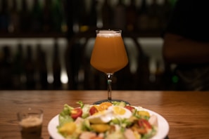 A tall glass of beer on a wooden table, accompanied by a salad on a white plate and a small shot glass with brown liquid. The background is dimly lit, with blurred reflections and shadows, suggesting a cozy and relaxed atmosphere.
