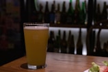 A glass of light-colored beer rests on a wooden table with a frothy head. The background features a dimly lit bar shelf filled with an assortment of various bottles, creating a cozy and intimate setting.