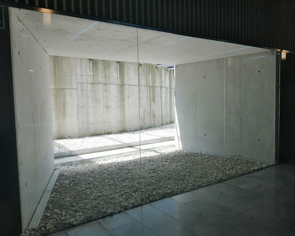 A minimalist architectural space with concrete walls and a large glass window. The area features a floor covered with small pebbles and has an open view towards a concrete wall outside, allowing natural light to filter in.