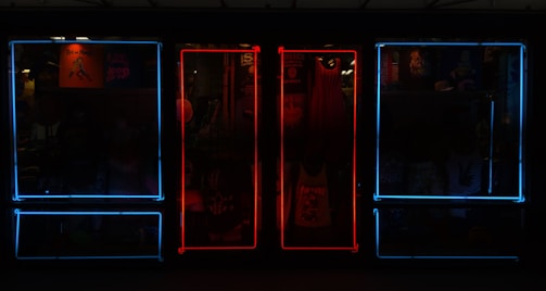 A storefront featuring vibrant neon lights, with a mixture of blue and red frames on windows. Inside the store, various items such as clothing and posters can be seen, adding depth and color to the scene.