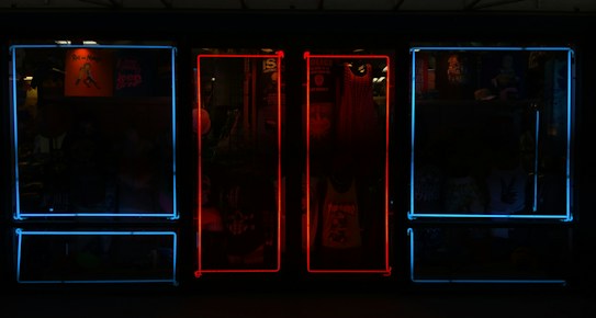 A storefront featuring vibrant neon lights, with a mixture of blue and red frames on windows. Inside the store, various items such as clothing and posters can be seen, adding depth and color to the scene.
