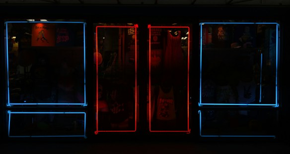 A storefront featuring vibrant neon lights, with a mixture of blue and red frames on windows. Inside the store, various items such as clothing and posters can be seen, adding depth and color to the scene.