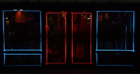 A storefront featuring vibrant neon lights, with a mixture of blue and red frames on windows. Inside the store, various items such as clothing and posters can be seen, adding depth and color to the scene.