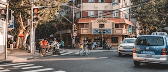 A busy urban intersection with cyclists, motorcycles, and cars. Pedestrians walk along the sidewalk lined with trees, and a multi-story building with a cafe is in the background. Traffic lights are visible, with some indicating red and others green.