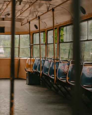 An empty and slightly worn-out interior of a vintage bus, featuring rows of blue and yellow seats. The windows are slightly foggy, and the lighting is soft, indicating an overcast day. Metal poles and a simple design suggest an older model of bus.