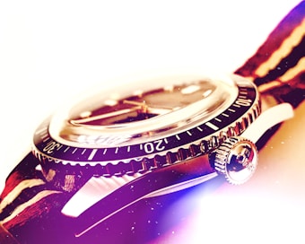 A close-up of a wristwatch featuring a detailed view of its bezel and crown. The watch has a metallic finish with clear numbered markings. The lighting creates a soft glow, highlighting the watch's polished surfaces and contributing to an overall vintage aesthetic.