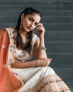 A person with braided hair sits on steps wearing traditional clothing adorned with intricate patterns. The outfit features a cream base with orange and gold detailing, and a matching dupatta. The person is accessorized with large earrings and a bracelet, and their pose is thoughtful with one hand near their face.