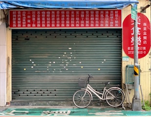A closed shop with a red and yellow signboard featuring Chinese characters above a green rolling shutter. A bicycle is parked in front of the shop next to a utility pole.